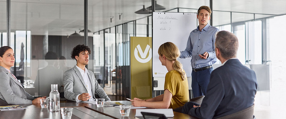 Mehrere Personen sitzen in einem modernen Besprechungsraum, während ein junger Mann eine Präsentation hält – Symbolbild für berufliche Neuorientierung, Karrierechancen und Weiterbildung beim Jobwechsel.